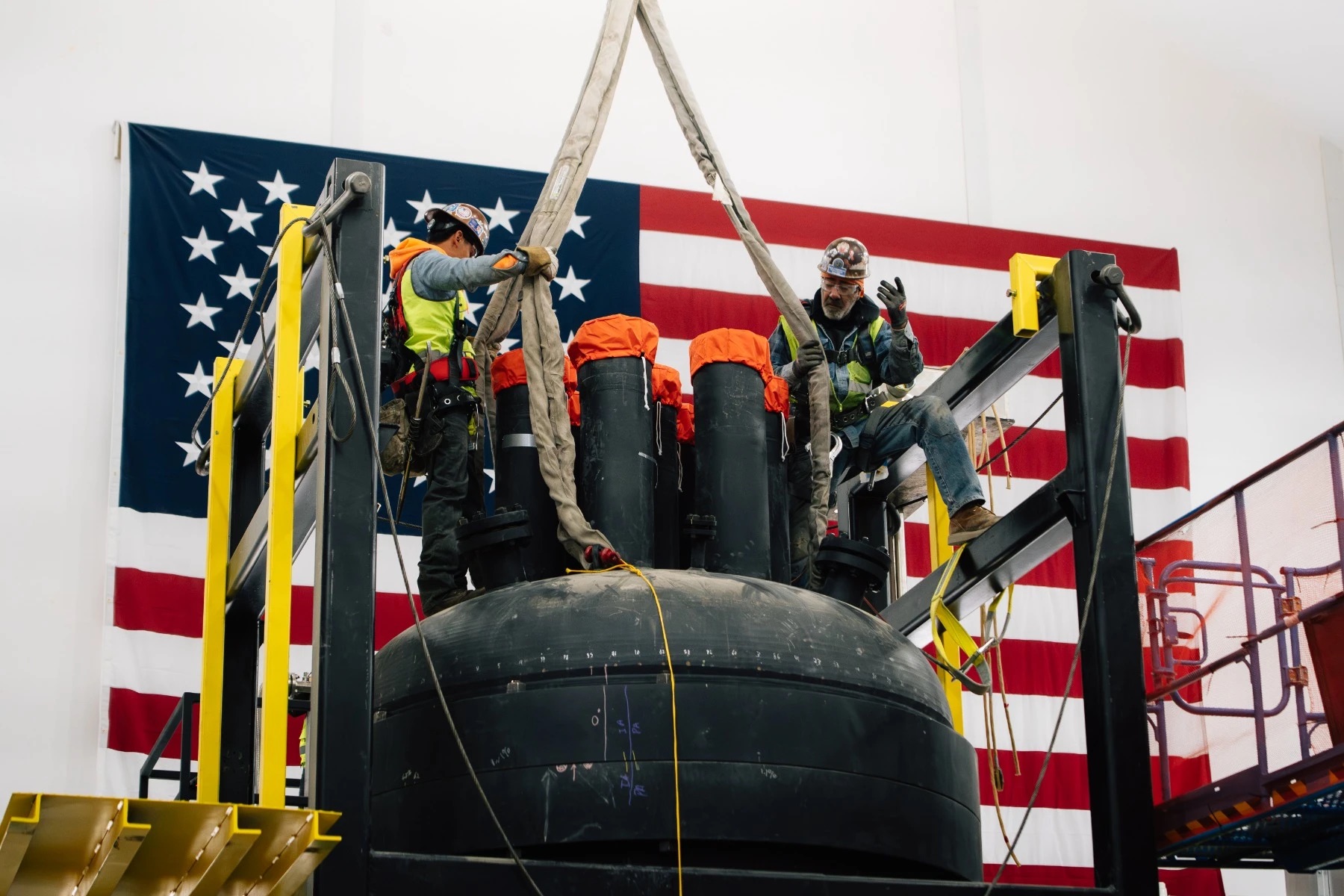 Workers rig a reactor vessel for lifting in front of an American flag